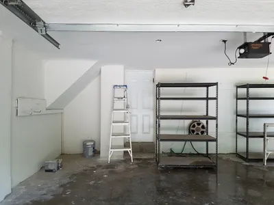 Interior view of a garage with newly painted white walls and a white textured ceiling. A white door, metal shelving units, a ladder, and a paint bucket are visible. The concrete floor is dirty, indicating recent work completion.